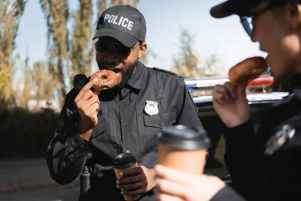 Policier afro-américain avec tasse en papier manger beignet avec collègue flou au premier plan à l'extérieur — Photo de stock