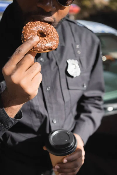 Cropped view of african american policeman with paper cup eating doughnut on blurred background outdoors — Stock Photo