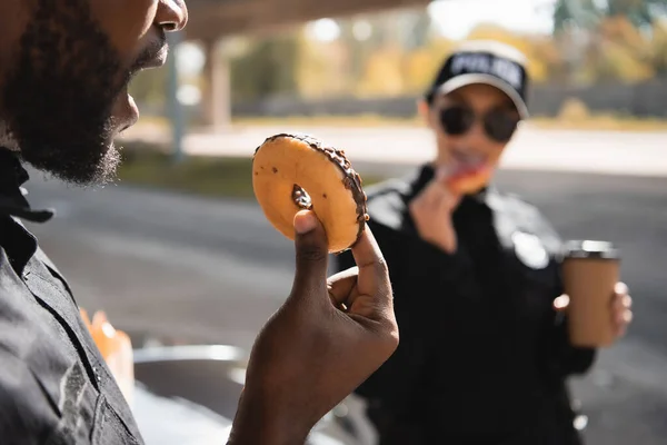 Vista de cerca del policía afroamericano con la boca abierta sosteniendo donut con compañero borroso en el fondo al aire libre - foto de stock