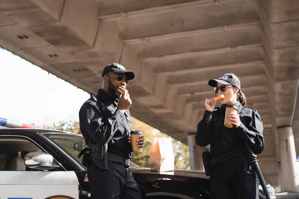 Multicultural police officers with paper cups eating doughnuts near patrol car on blurred background on urban street — Stock Photo
