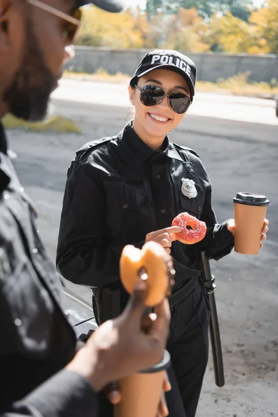 Happy policewoman with doughnut and paper cup standing near african american colleague on blurred foreground outdoors — Stock Photo