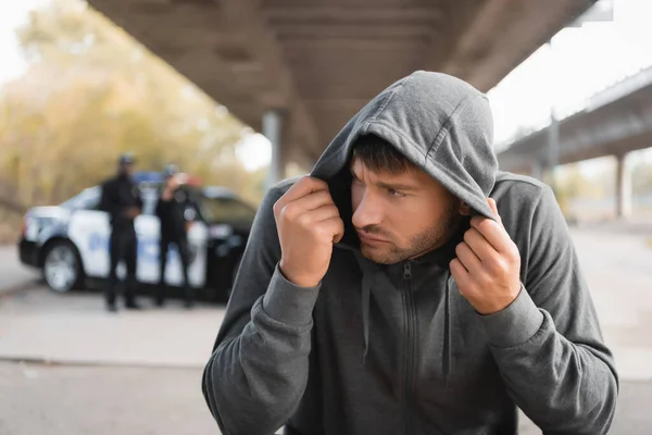 Assustado encapuzado ofensor escondendo com policiais desfocados no fundo ao ar livre — Fotografia de Stock