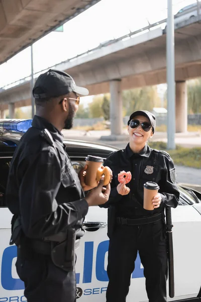 Сотрудники многонациональной полиции с бумажными стаканчиками и пончиками разговаривают возле патрульной машины на размытом фоне на городской улице — стоковое фото