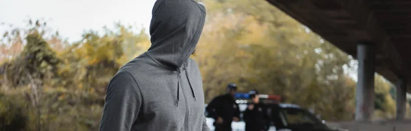 Преступник с капюшоном, смотрящий в сторону с размытыми полицией на заднем плане, баннер — стоковое фото