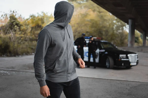 Hooded thief looking away with blurred multicultural police officers on background outdoors — Stock Photo