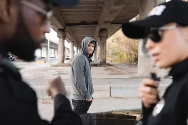 Délinquant à capuchon insatisfait regardant des policiers multiculturels flous au premier plan dans la rue urbaine — Photo de stock