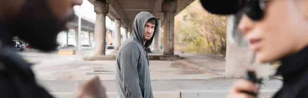 Displeased hooded offender looking at blurred multicultural police officers on foreground outdoors, banner — Stock Photo