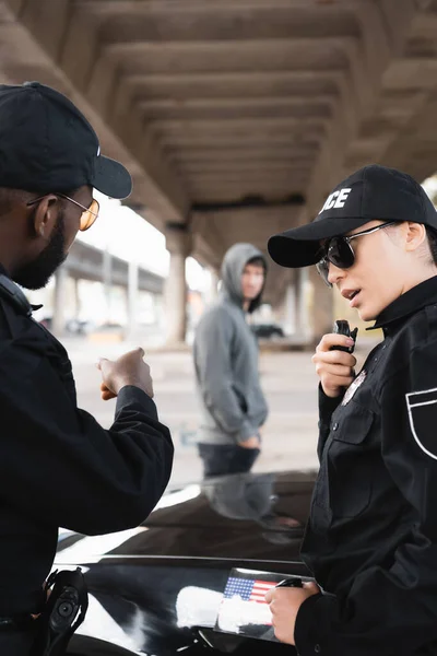 Policewoman talking on radio set near african american colleague pointing with finger at blurred offender on background outdoors — Stock Photo