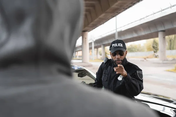 Серьезный африканский американский полицейский, указывающий пальцем на размытого преступника в капюшоне на переднем плане на городской улице — стоковое фото