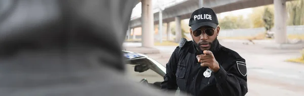 Grave agent de police afro-américain pointant du doigt délinquant cagoulé flou au premier plan dans la rue urbaine, bannière — Photo de stock