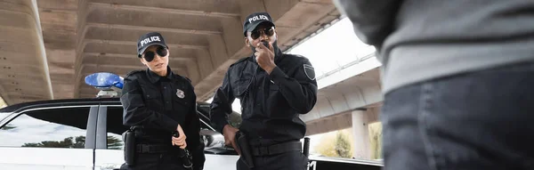 Afrikanisch-amerikanischer Polizeibeamter spricht am Funkgerät in der Nähe eines Kollegen, der einen Polizeiknüppel zückt, mit verwischtem Täter im Vordergrund, Banner — Stockfoto