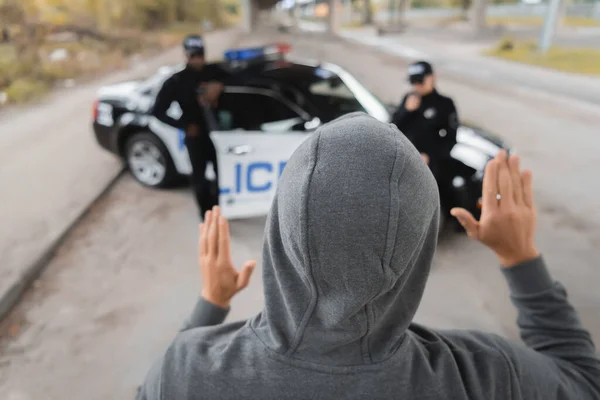 Vista trasera del delincuente encapuchado mostrando las manos con oficiales de policía multiculturales borrosas en el fondo al aire libre - foto de stock