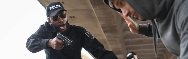 Policial americano africano agressivo gritando enquanto apontando por arma para o infrator rendido em primeiro plano desfocado ao ar livre, banner — Fotografia de Stock