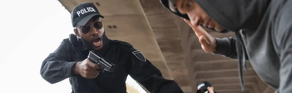 Aggressive african american policeman shouting while aiming by gun at surrendered offender on blurred foreground outdoors, banner — Stock Photo