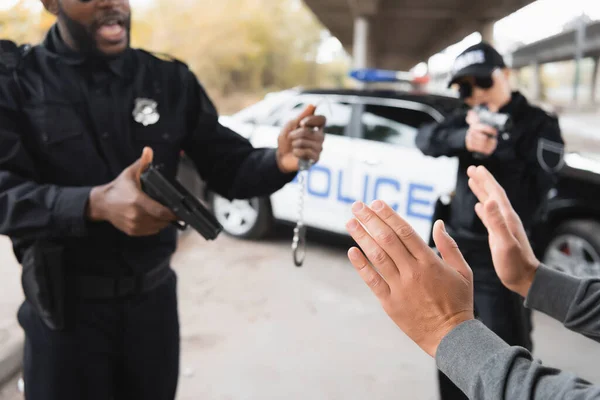 Delincuente mostrando las manos mientras agentes de policía multiculturales sosteniendo pistolas y esposas en el fondo borroso al aire libre - foto de stock