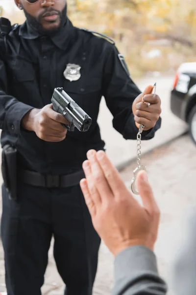 Visão recortada do policial afro-americano com algemas apontando por arma para ofensor desfocado em primeiro plano ao ar livre — Fotografia de Stock