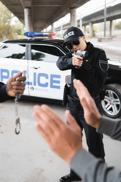 Policewoman aiming by pistol at surrendered offender with blurred african american colleague on foreground outdoors — Stock Photo