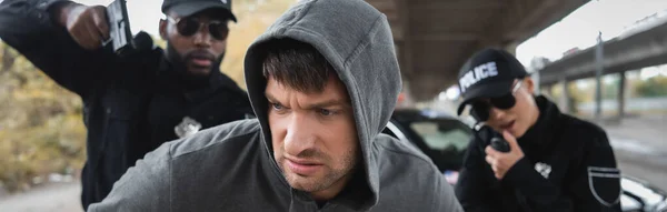 Ladrón disgustado mirando hacia otro lado cerca de policías multiculturales apuntando con pistolas sobre fondo borroso al aire libre, pancarta - foto de stock