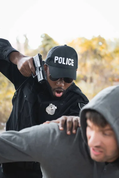 Aggressive african american policeman aiming with pistol while arresting hooded offender on blurred foreground outdoors — Stock Photo