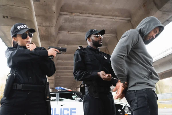 African american policeman handcuffing offender while colleague aiming with pistol near patrol car on urban street — Stock Photo