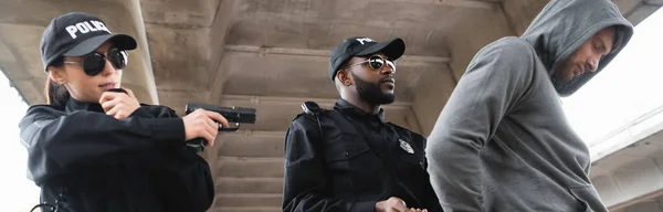 Африканский американский полицейский арестовывает преступника в капюшоне рядом с коллегой, стреляющим из пистолета на улице, баннер — стоковое фото