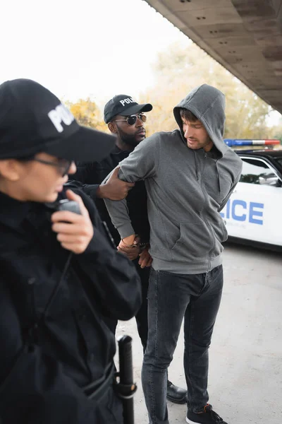 African american policeman handcuffing hooded offender with blurred colleague on foreground outdoors — Stock Photo