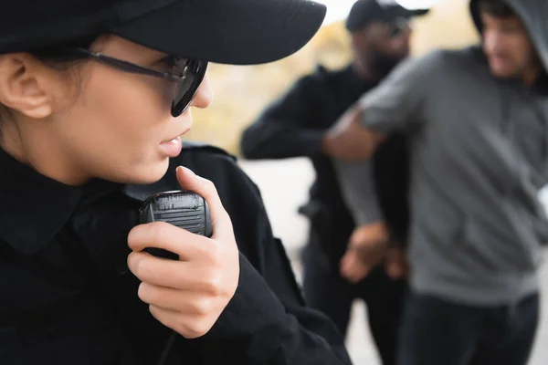 Close up view of policewoman talking on radio set during african american colleague arresting offender on blurred background — Stock Photo