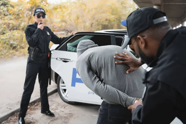 Американский полицейский африканского происхождения надел наручники на преступника рядом с коллегой, разговаривающим по радио возле патрульной машины на размытом фоне — стоковое фото