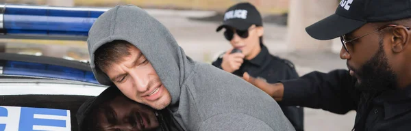 African american policeman arresting hooded offender near patrol car with blurred colleague on background outdoors, banner — Stock Photo