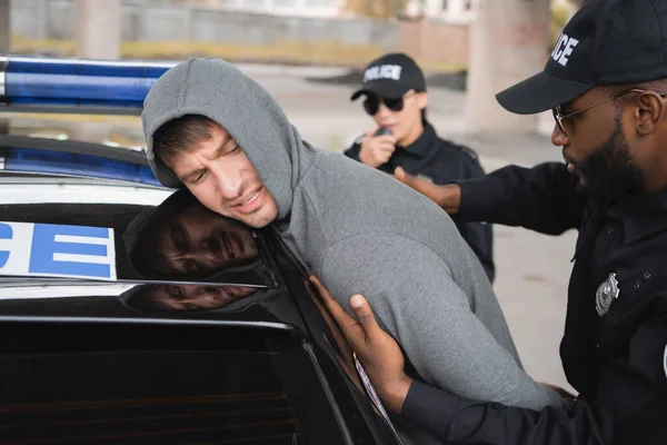 Africano americano policial frisking irritado encapuzado infrator inclinando-se em carro de patrulha no fundo borrado ao ar livre — Fotografia de Stock