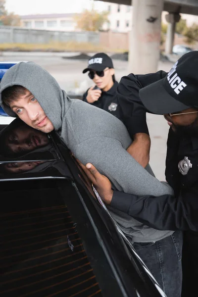 Afroamericano policía registrando triste con capucha delincuente apoyado en el coche sobre fondo borroso al aire libre - foto de stock