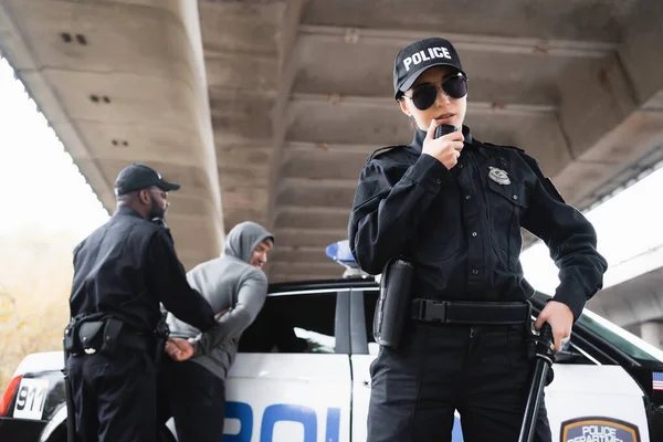 Policewoman in sunglasses talking on radio set near african american colleague arresting offender on blurred background — Stock Photo