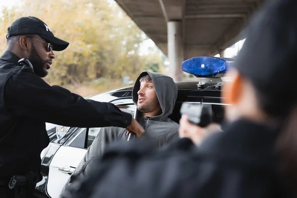 Policial afro-americano com truncheon prendendo criminoso encapuzado com colega embaçado apontando por arma em primeiro plano ao ar livre — Fotografia de Stock