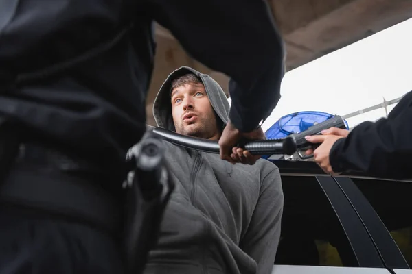 Hooded offender looking at african american policeman with truncheon near policewoman aiming by pistol on blurred foreground outdoors — Stock Photo