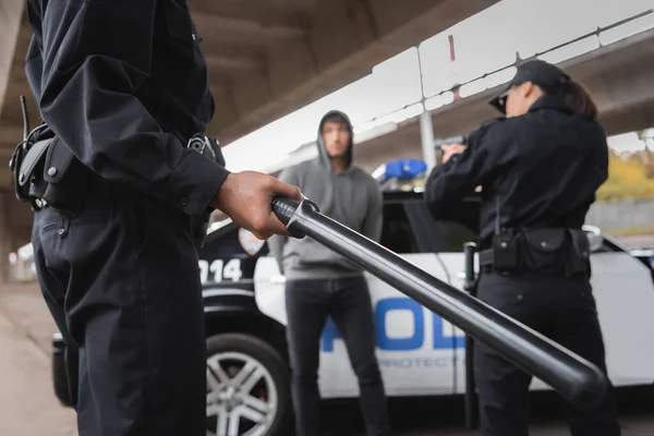 Afro-americano policial segurando truncheon perto colega apontando com pistola e ofensor no fundo borrado ao ar livre — Fotografia de Stock