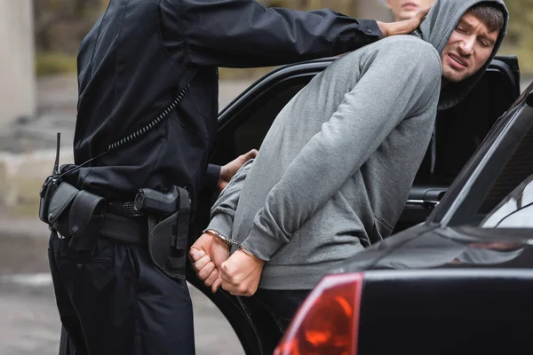 Un policier afro-américain arrête un délinquant à capuchon en colère près d'une voiture de patrouille sur fond flou à l'extérieur — Photo de stock