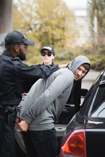 Un policier afro-américain arrête un délinquant à capuchon en colère près d'un collègue et d'une voiture de patrouille sur fond flou à l'extérieur — Photo de stock