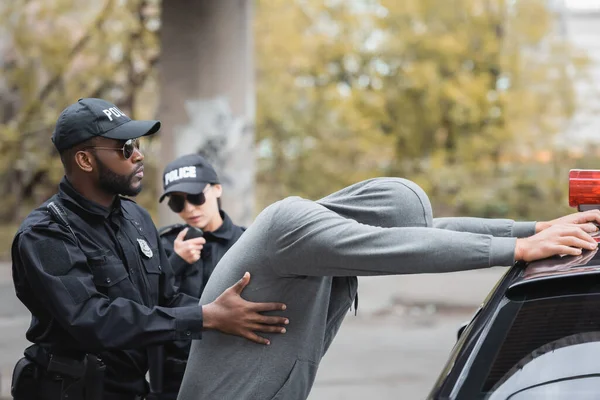 Africano americano poliziotto perquisire incappucciato delinquente appoggiato su pattuglia auto vicino collega su sfondo sfocato all'aperto — Foto stock