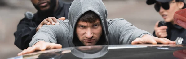 Angry offender with outstretched hands leaning on patrol car with blurred multicultural police officers on background, banner — Stock Photo