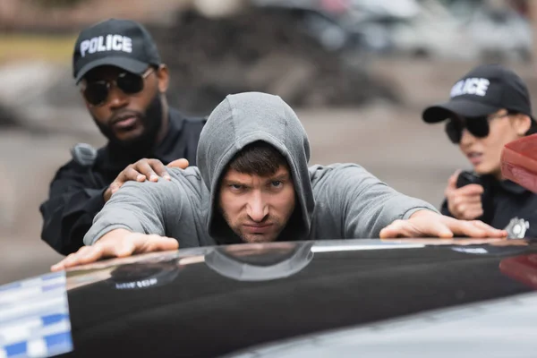 Ofensor enojado con las manos extendidas apoyadas en el coche patrulla durante el registro con oficiales de policía multiculturales borrosas en el fondo - foto de stock