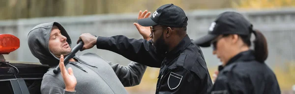 African american policeman with truncheon arresting hooded offender with blurred colleague on foreground outdoors, banner — Stock Photo