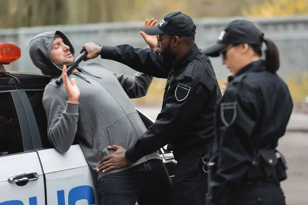 Africano policial americano com truncheon flertando ofensor com as mãos levantadas em primeiro plano borrado ao ar livre — Fotografia de Stock