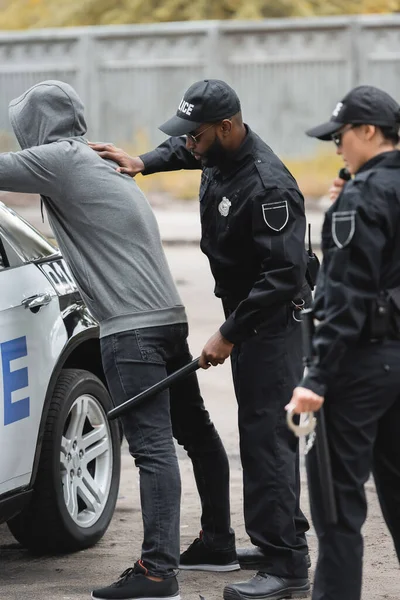 Policía afroamericano con truncheon registrando delincuente encapuchado cerca de patrulla coche con compañero borroso en primer plano al aire libre - foto de stock