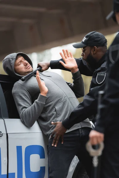 Poliziotto afroamericano con truncheon perquisire criminale incappucciato con le mani alzate su primo piano sfocato all'aperto — Foto stock