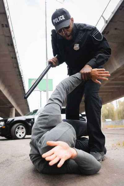 Vista basso angolo di agente di polizia aggressivo con manganello arresto criminale incappucciato sdraiato sulla strada urbana — Foto stock