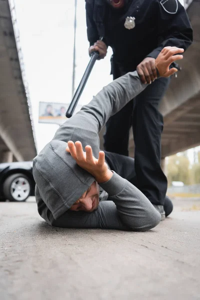 African american police officer with truncheon arresting hooded thief lying on street on blurred background — Stock Photo