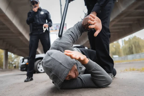 African american policeman with truncheon arresting hooded offender covering face while lying on street on blurred background — Stock Photo