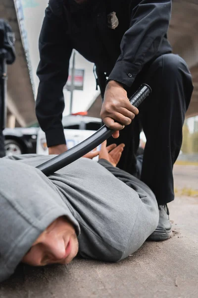 Policial afro-americano com truncheon prendendo ladrão encapuzado deitado na rua em fundo turvo — Fotografia de Stock
