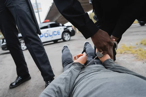 Vista recortada del policía afroamericano esposando a delincuente encapuchado acostado en la calle sobre fondo borroso al aire libre - foto de stock