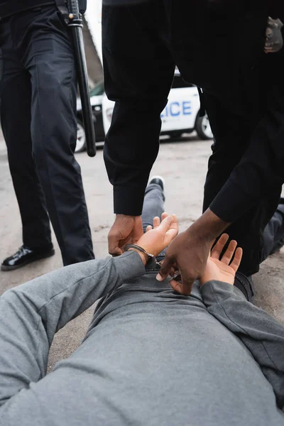 Vista cortada do policial afro-americano algemando infrator deitado na rua perto colega no fundo borrado ao ar livre — Fotografia de Stock